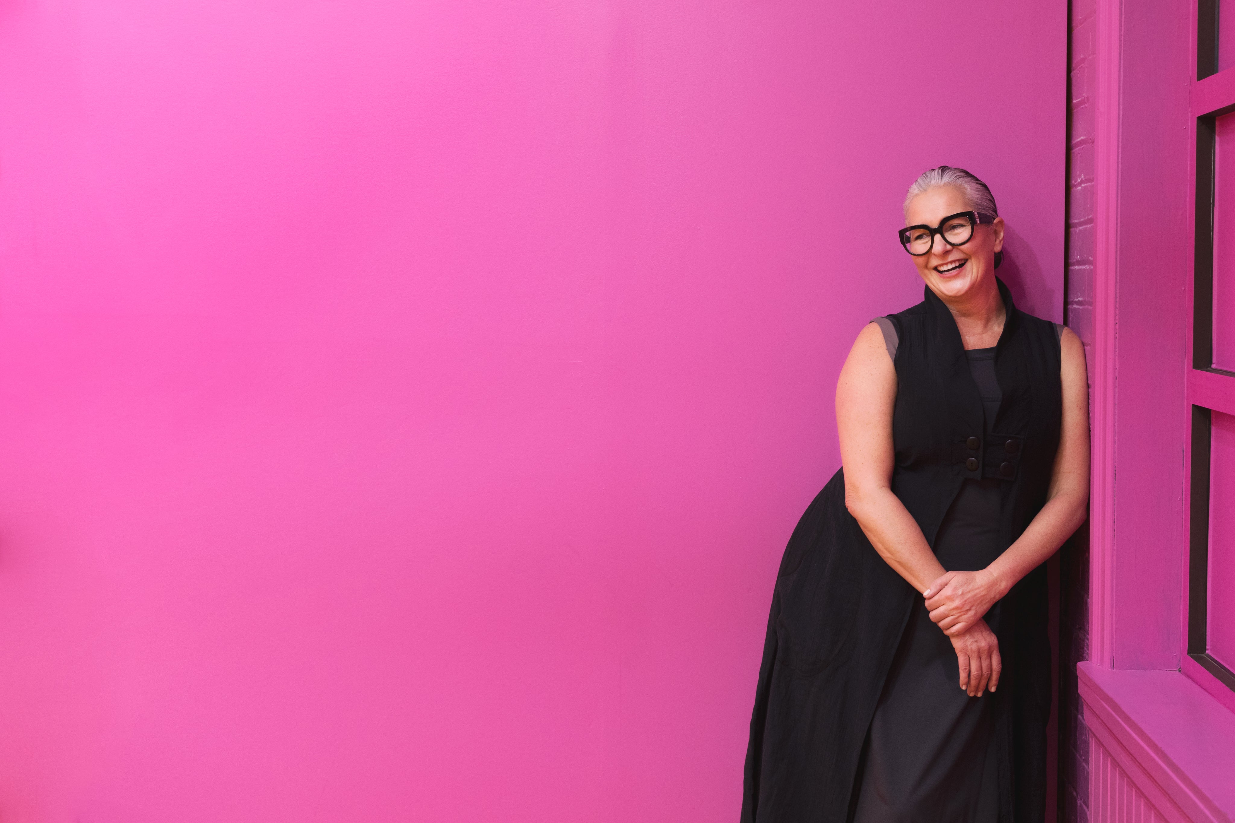 Confident woman in black outfit smiling against pink wall representing Averon modern fashion style.